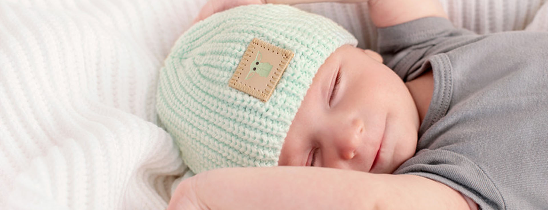 A sleeping baby wearing a light green knit beanie with a small patch on the front featuring Grogu from Star Wars: The Mandalorian