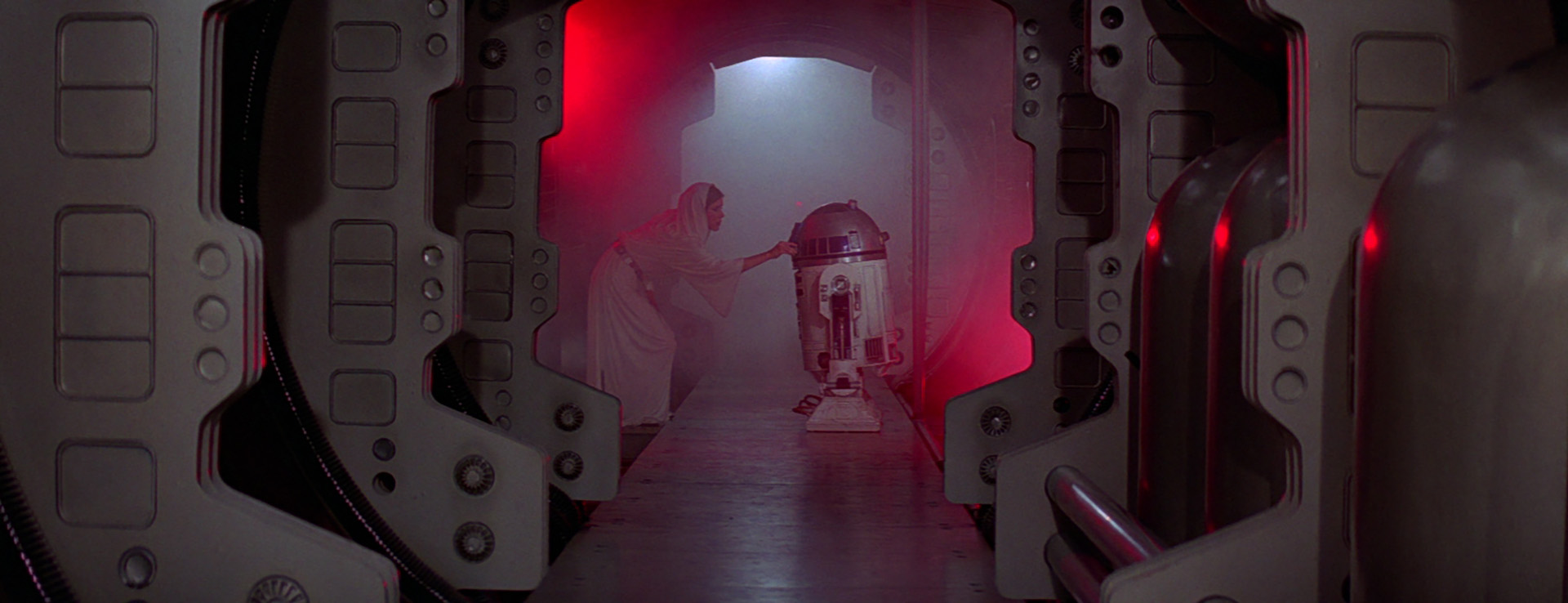 A film still from Star Wars: A New Hope. At the end of a hallway, Princess Leia has her hand on R2-D2.