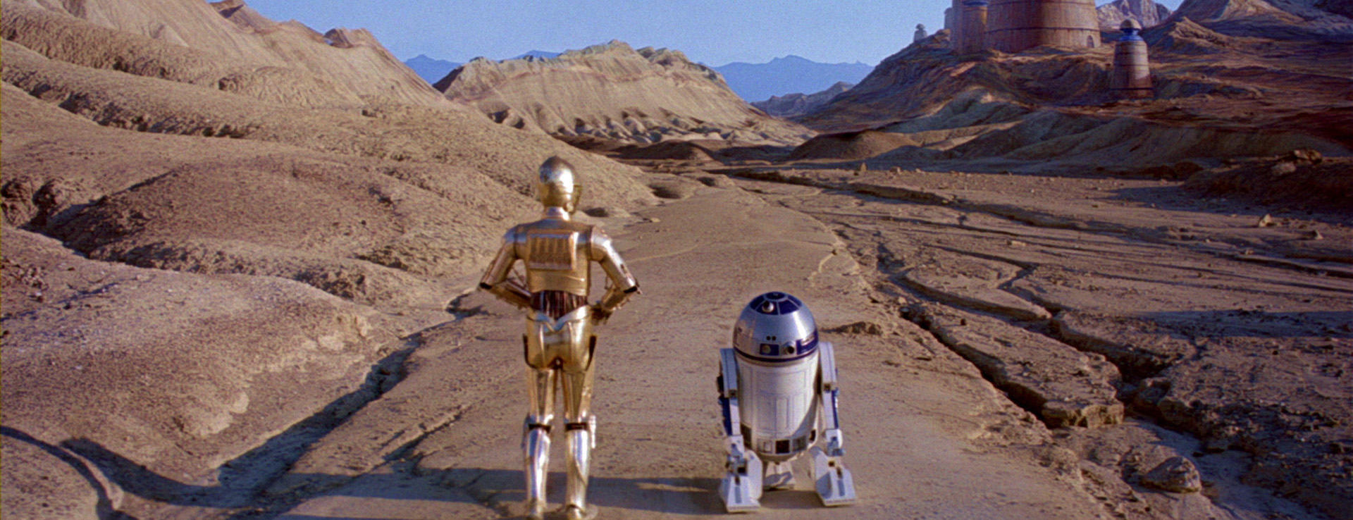 A Star Wars film still featuring C3P-O and R2-D2 walking side by side down a desert path.