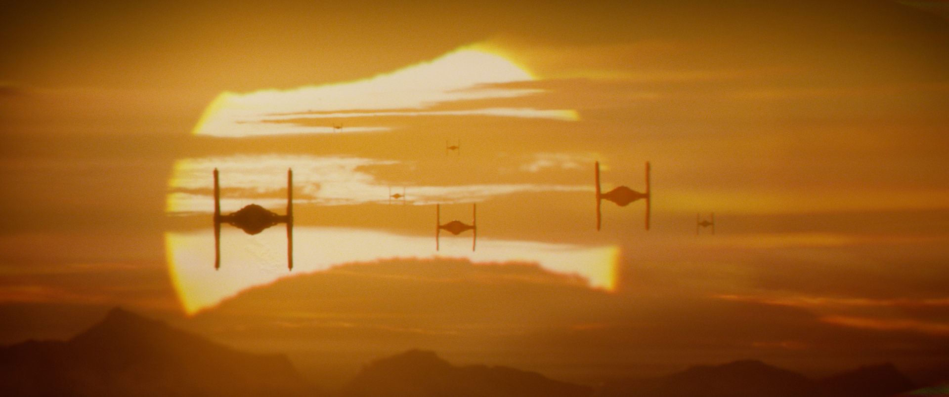 A Star Wars film still featuring three TIE fighters approaching in front of a setting sun.