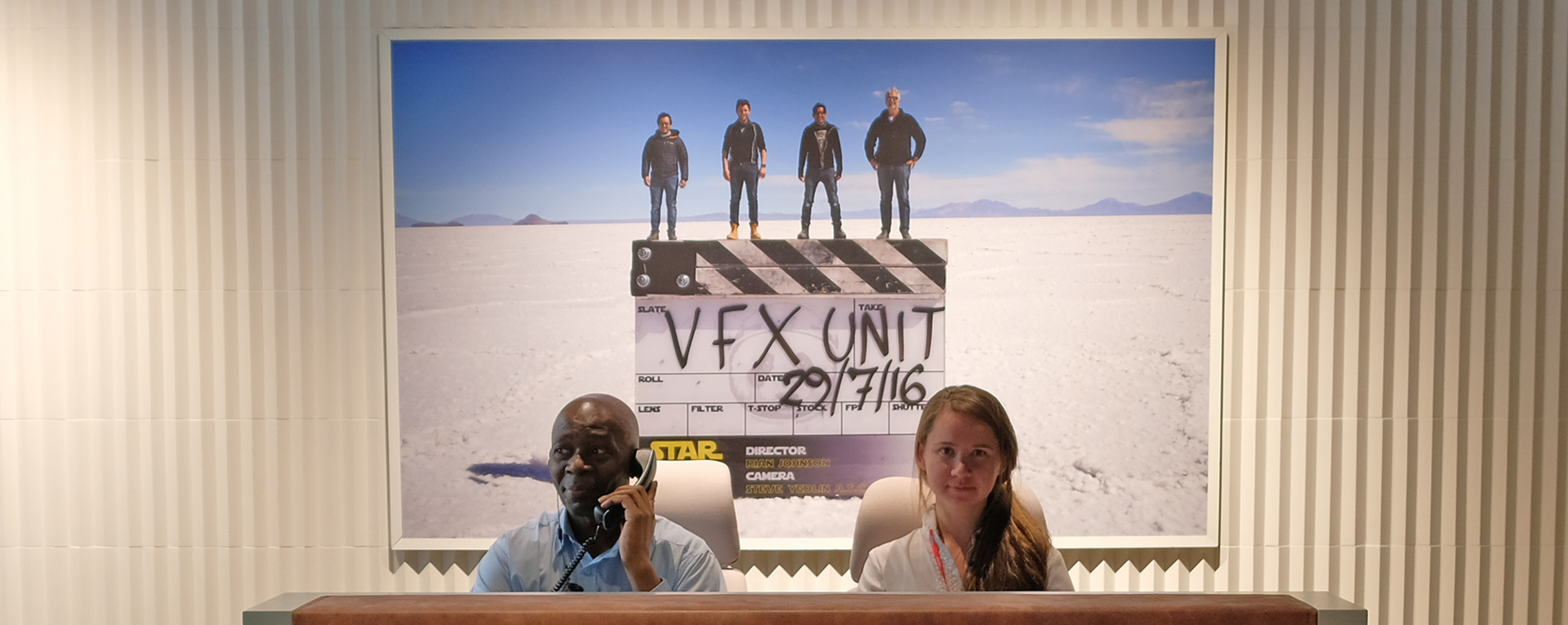 The public lobby at Industrial Light & Magic's London Studio. Two employees sit at a front desk under a large piece of art featuring a VFX unit clapperboard.