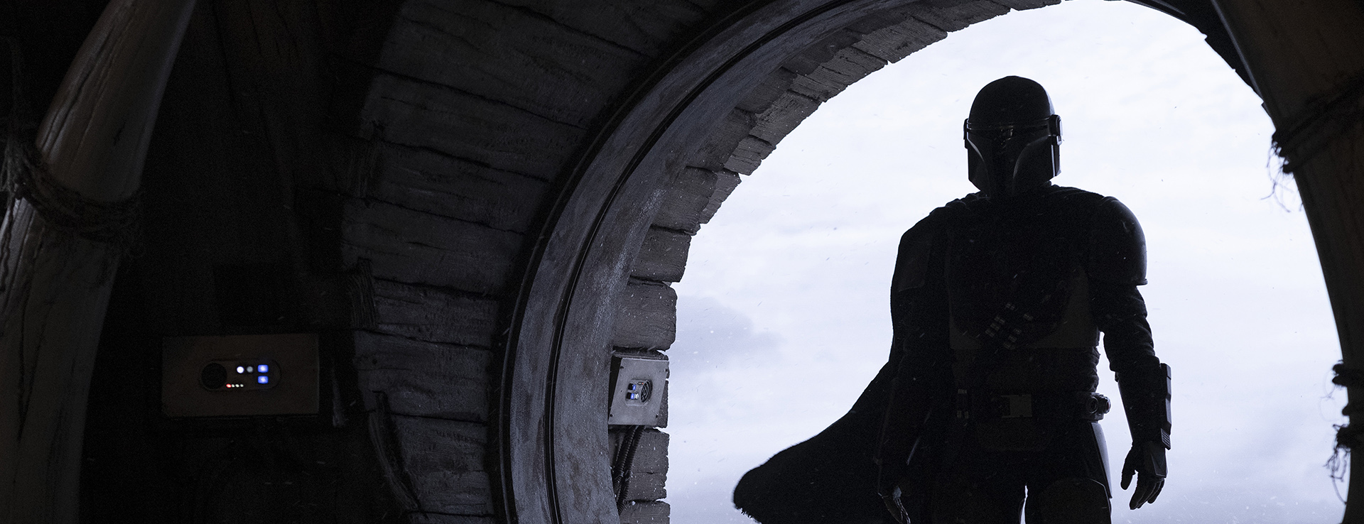 A still from Star Wars: The Mandalorian. The Mandalorian stands in front of circular open doorway.