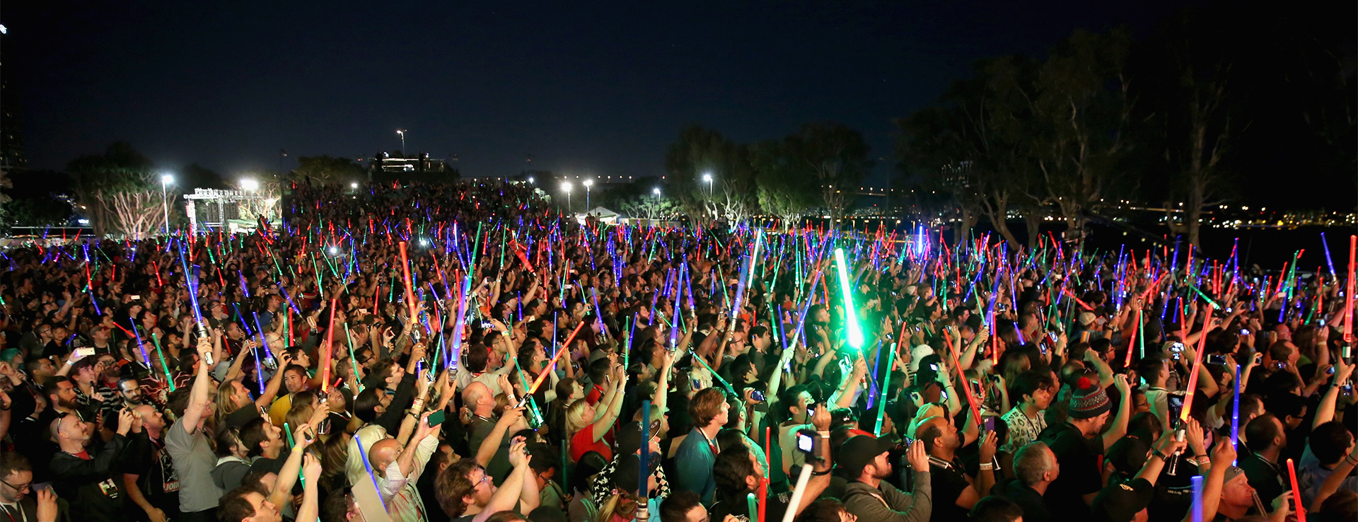 A large group of fans at Star Wars Celebration hold up their lightsabers into the night sky.