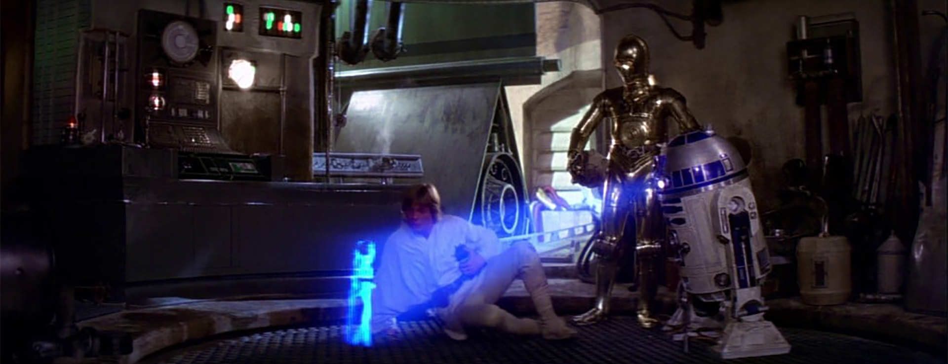 A film still from Star Wars: A New Hope. Luke and C3P-O look on as R2-D2 shows them a hologram message from Princess Leia.