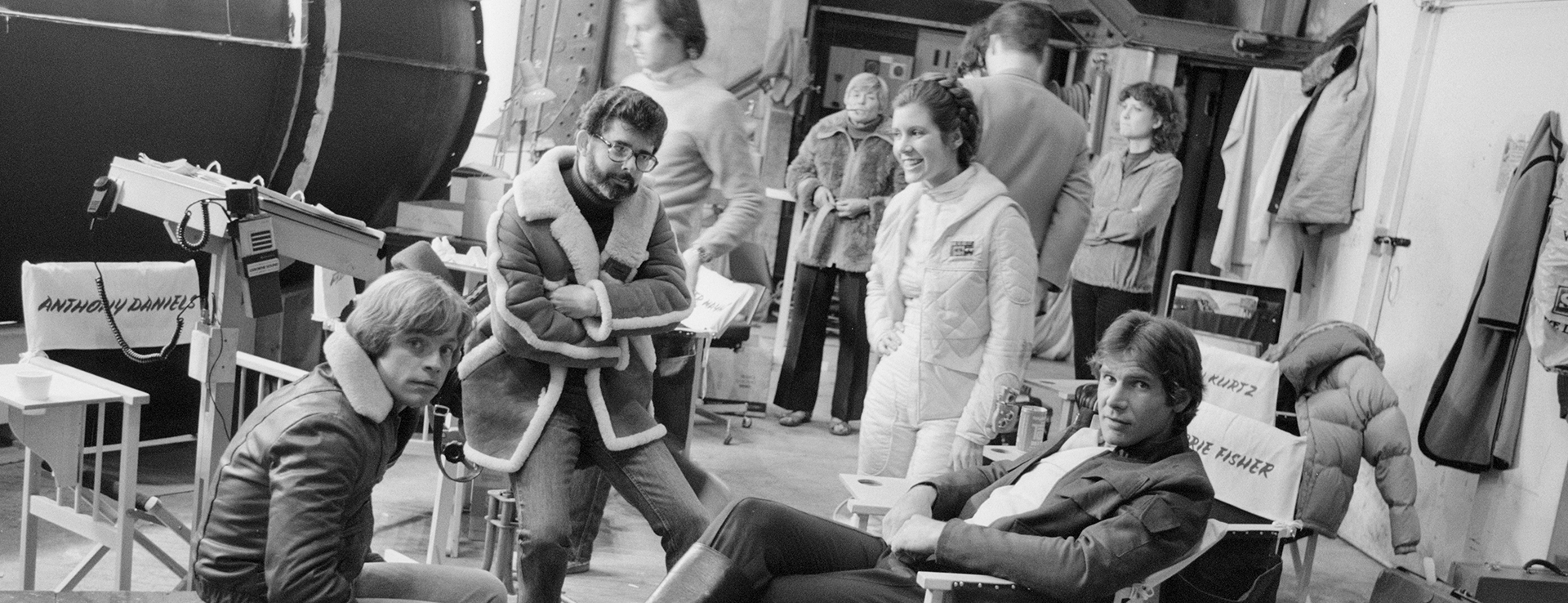 A black and white behind the scenes photo from Star Wars: The Empire Strikes Back featuring George Lucas, Carrie Fisher, Mark Hamill and Harrison Ford, with some crew members in the background