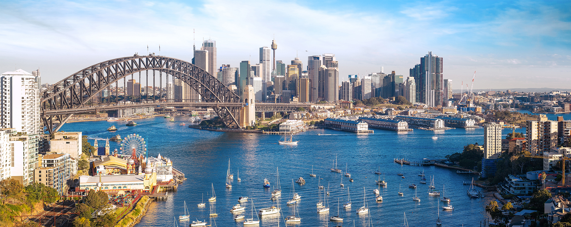 A wide view of Sydney Harbour. There are sailboats in the Harbour, a view of Sydney Harbour Bridge, and the Sydney skyline in the background.